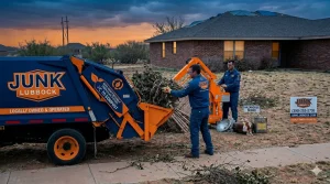 Two men in Junk Lubbock branded work shirts actively load extensive woody yard debris and broken branches into the hydraulic hopper of a large white garbage truck under a dramatic, blue and orange-toned twilight sky
